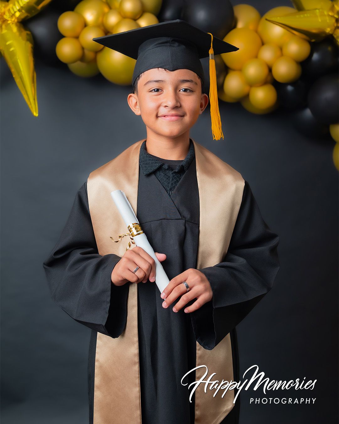 Graduate in traditional cap and gown with gold balloons and black backdrop.