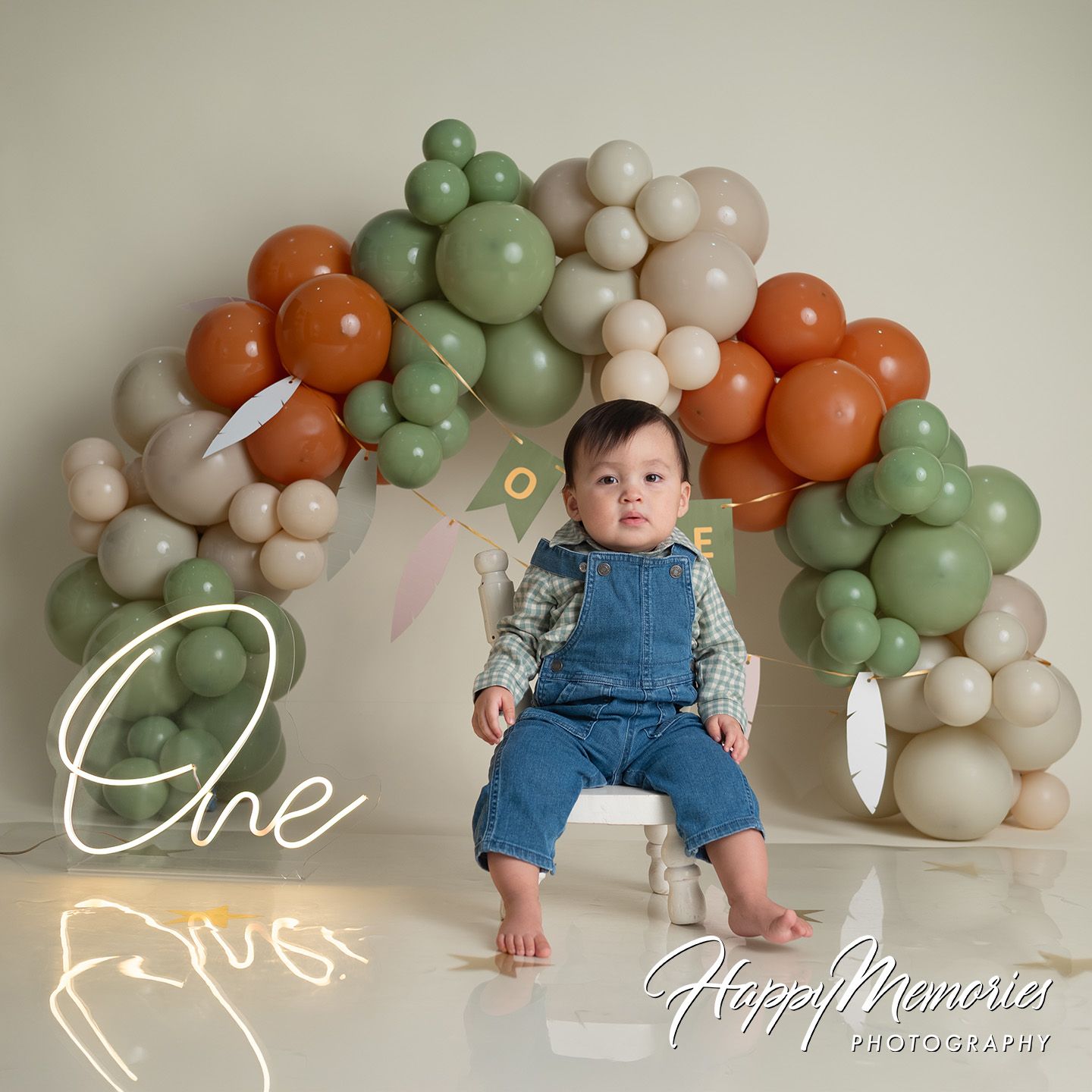 Baby's first birthday photo with colorful balloon arch backdrop, infant sitting on white cube, wearing denim overalls.