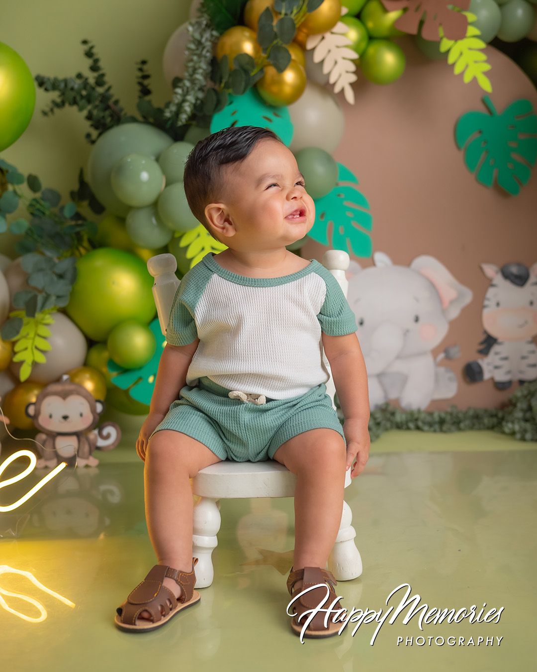 A baby in a teal outfit sits among a festive green and gold balloon arch with tropical leaf decorations.