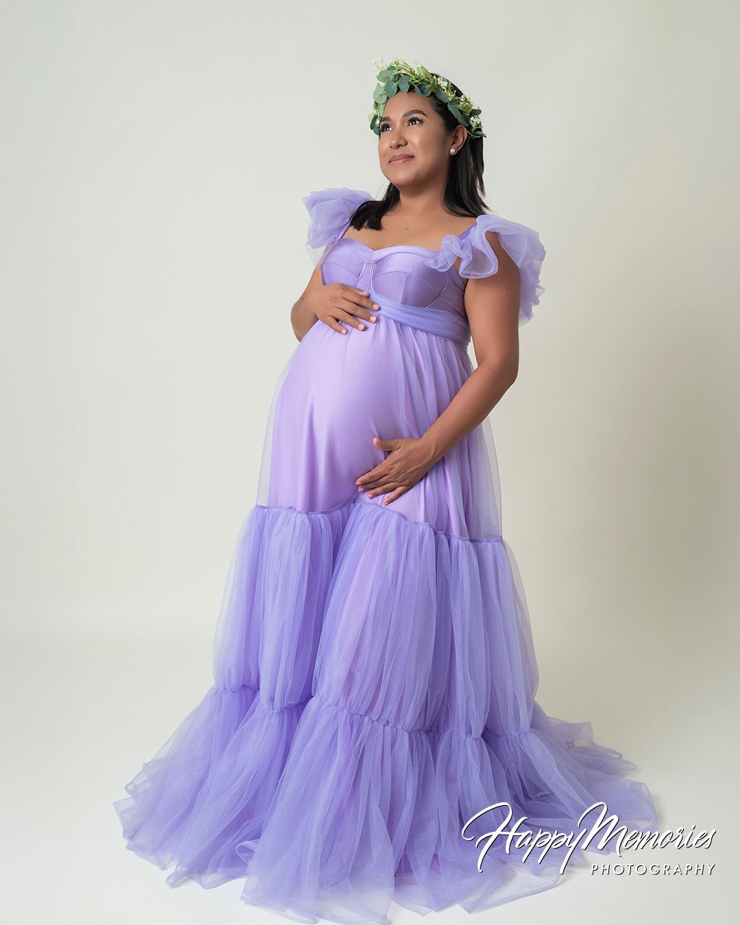 Maternity portrait in lavender tulle gown with floral crown against white backdrop.