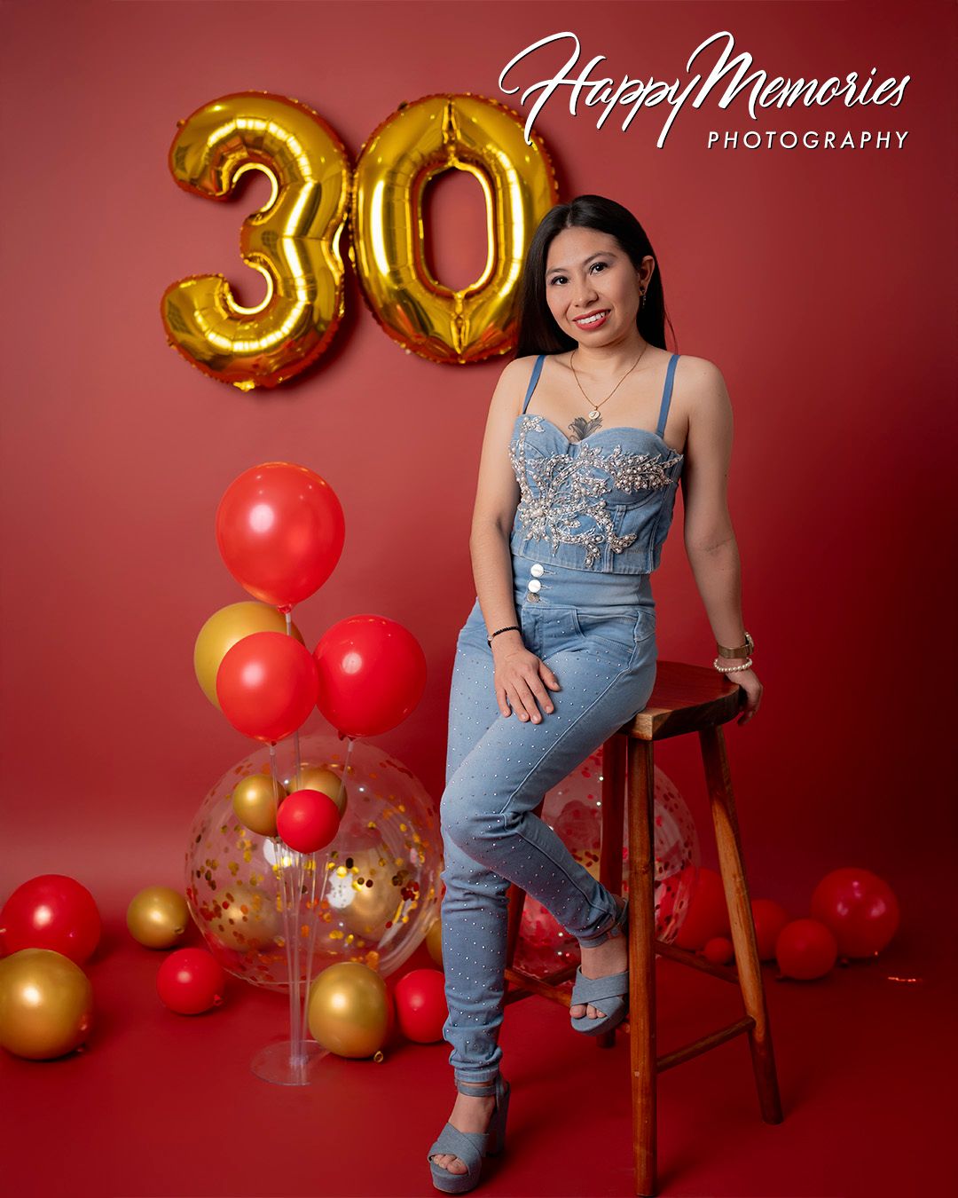 Birthday portrait session with denim outfit and red and gold balloon decorations against burgundy background.