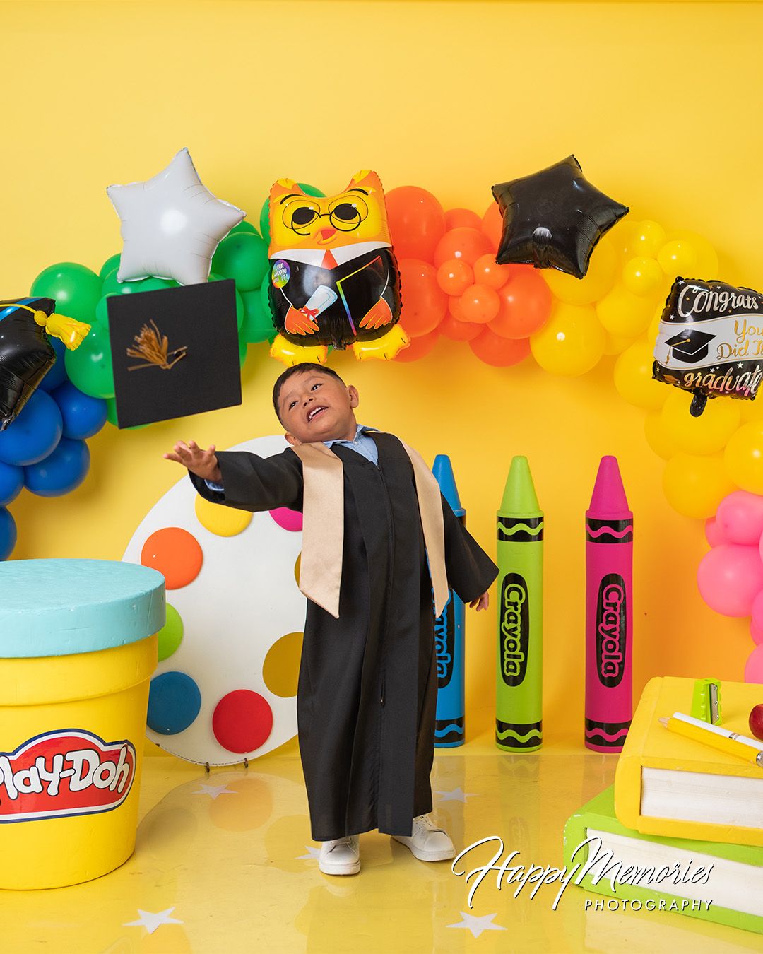 Colorful graduation themed photo setup with Play-Doh containers, crayons, and balloons against yellow backdrop.