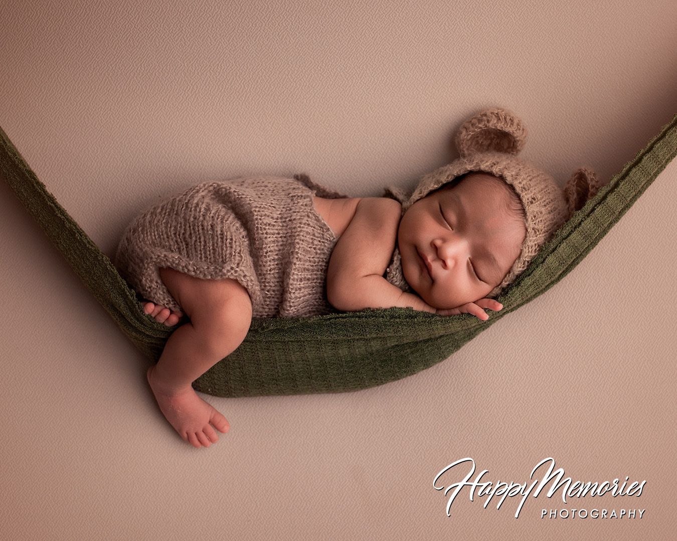 A sleeping newborn baby wrapped in brown knit fabric lies peacefully in a hanging hammock during a professional photo session.