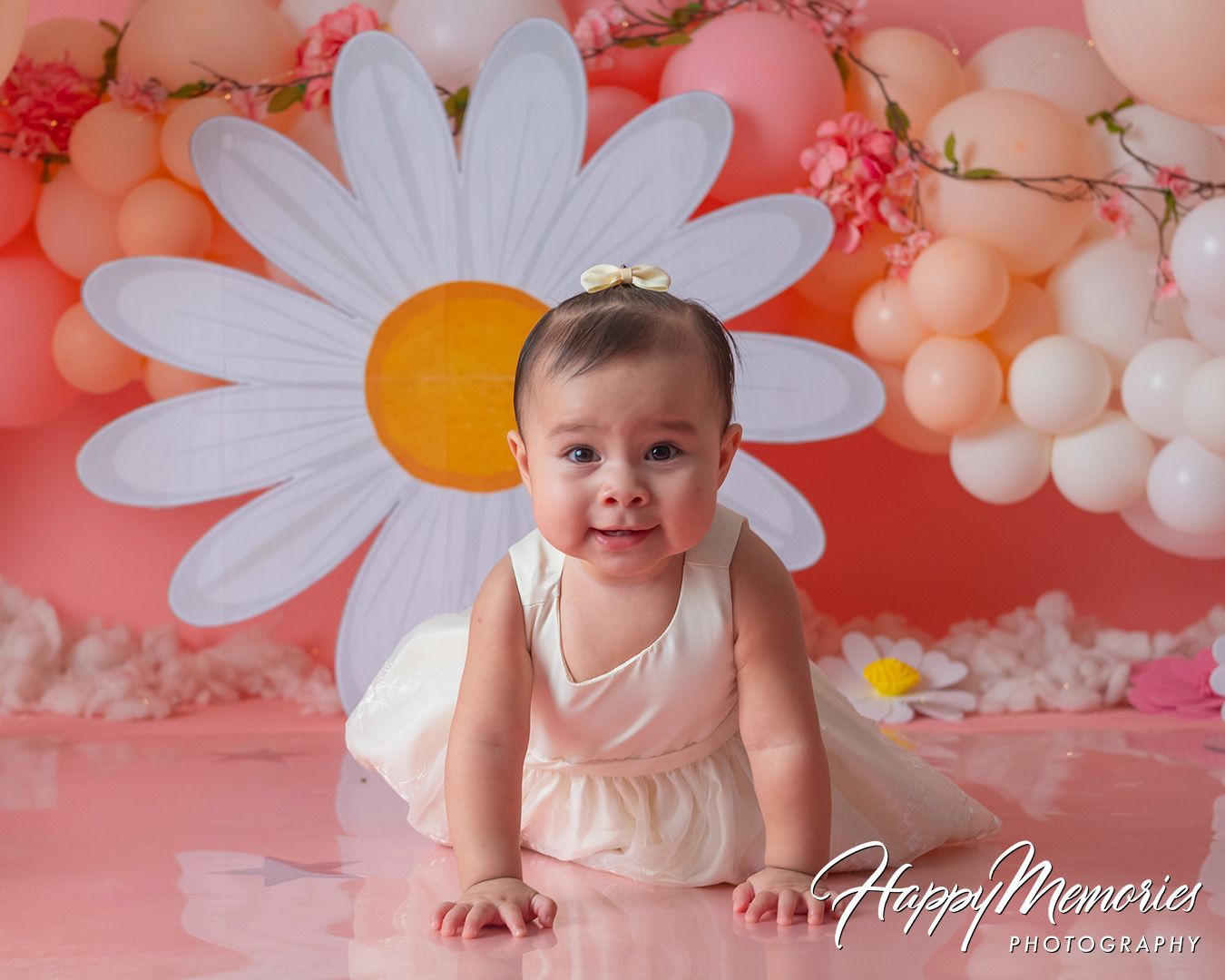 Baby in pink outfit sits against coral backdrop with white daisy decorations and pink balloon garland.