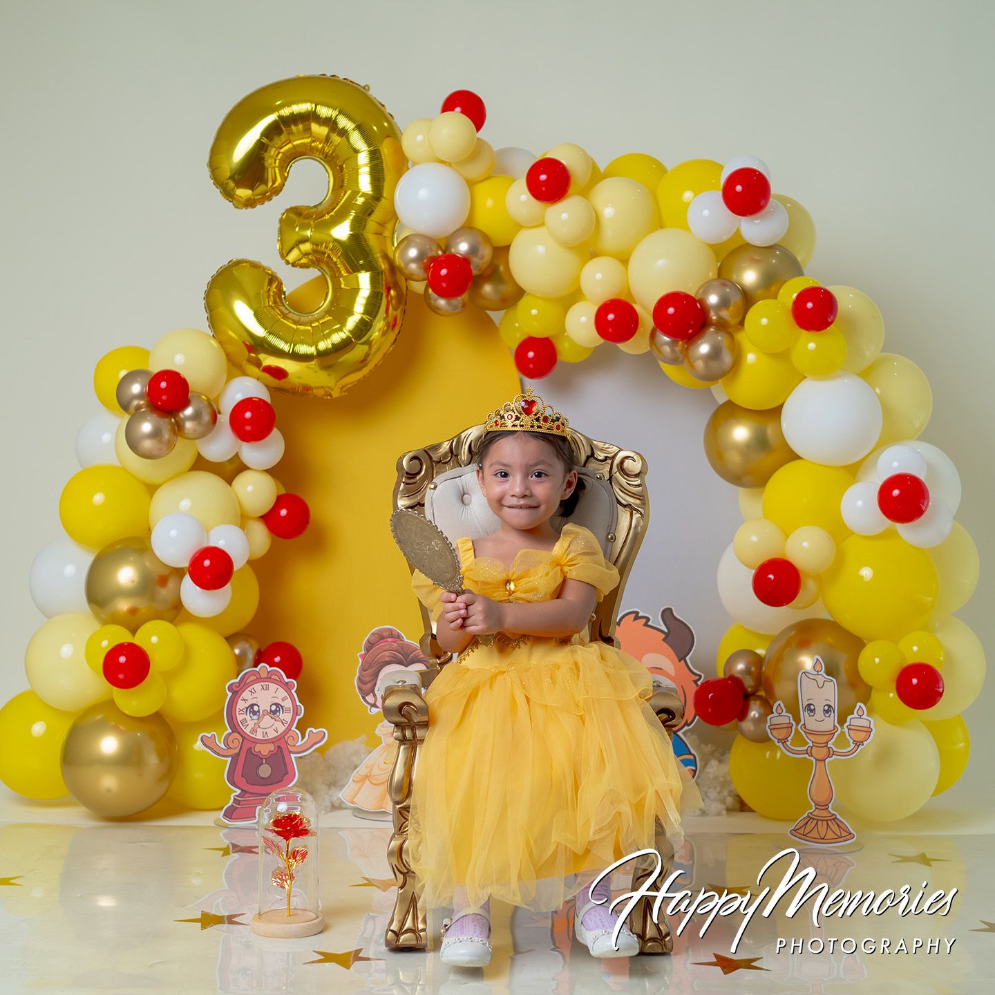 A toddler in a yellow Belle dress celebrates her 3rd birthday with a Beauty and the Beast balloon arch backdrop.