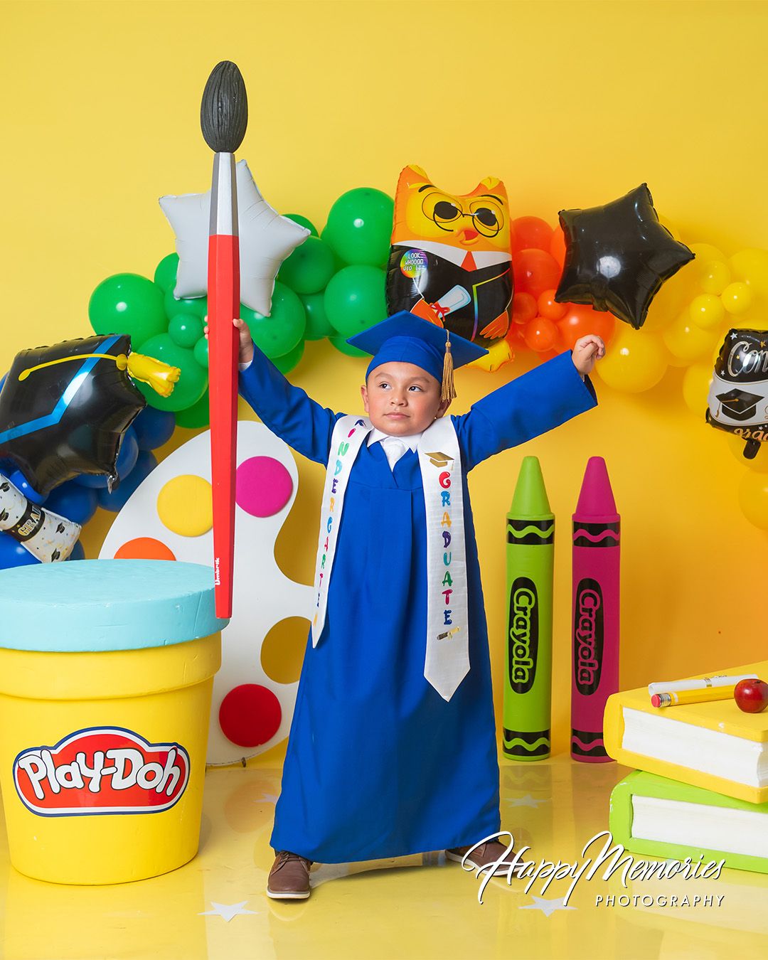 Creative graduation photo display featuring oversized crayons, Play-Doh, and colorful balloon decorations.