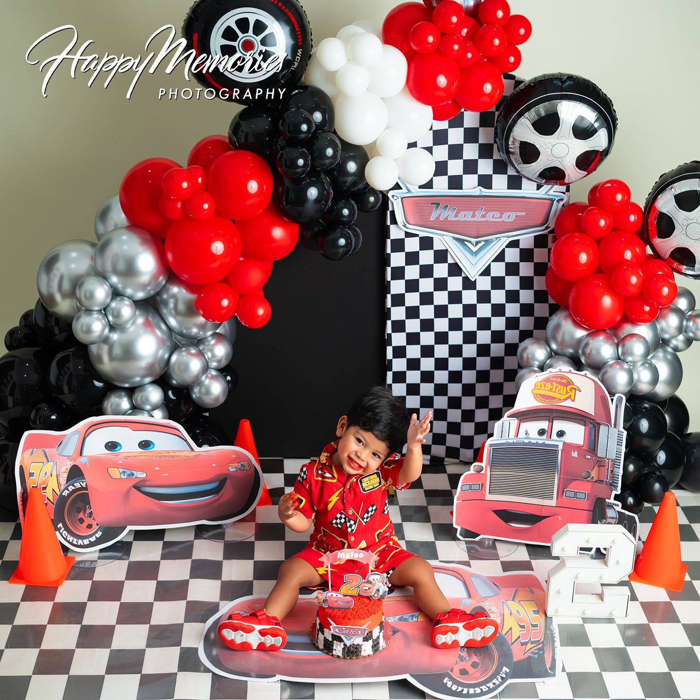 A toddler celebrates a Cars-themed birthday with Lightning McQueen cutouts, checkered backdrop, and red, black, silver balloons.