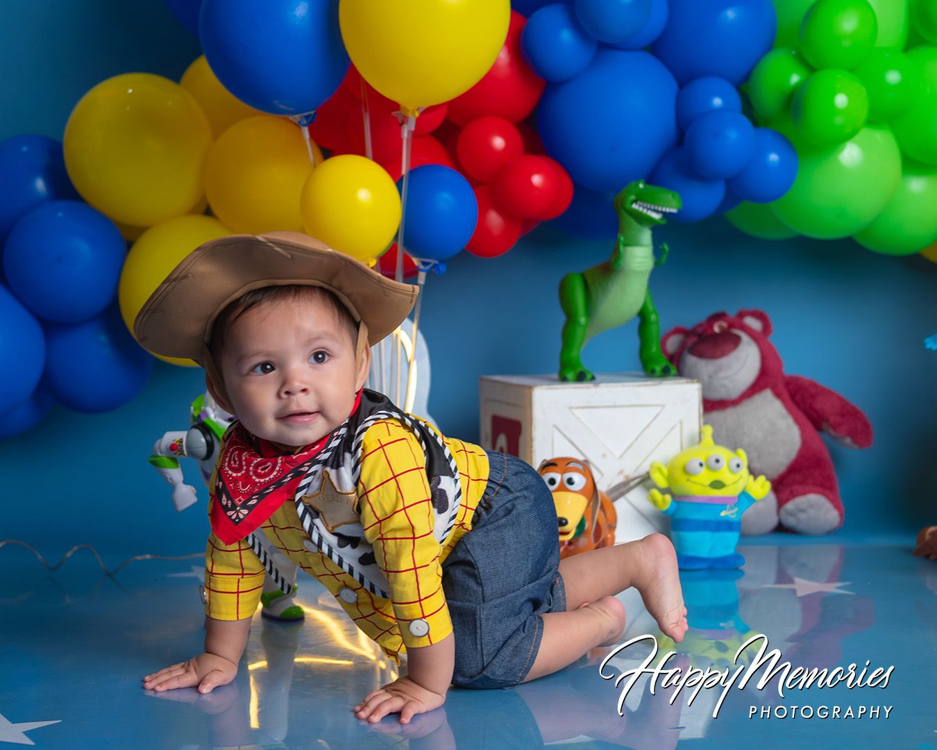 Toy Story themed birthday photo setup with colorful balloon backdrop, cowboy outfit, and dinosaur decorations.