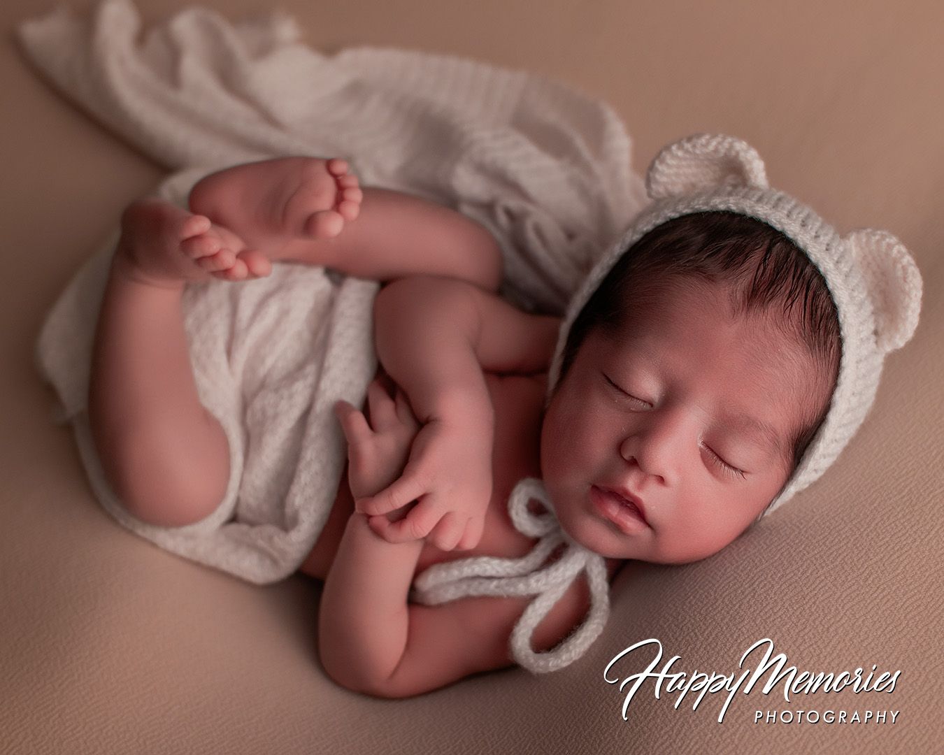 Sleeping newborn baby wearing white bear ear bonnet lying on soft beige blanket during professional photoshoot.