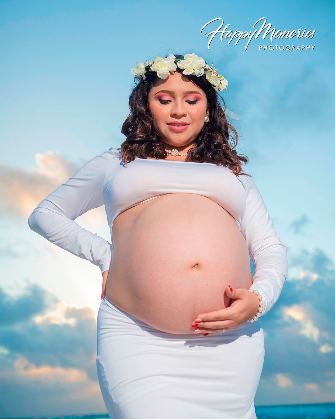 Beachside maternity portrait in white dress with floral crown at sunset.