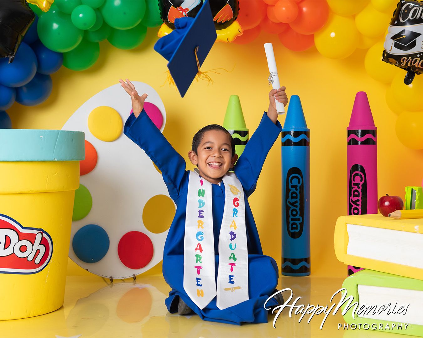 Colorful graduation photo setup with Play-Doh containers and crayon decorations against a bright yellow background.