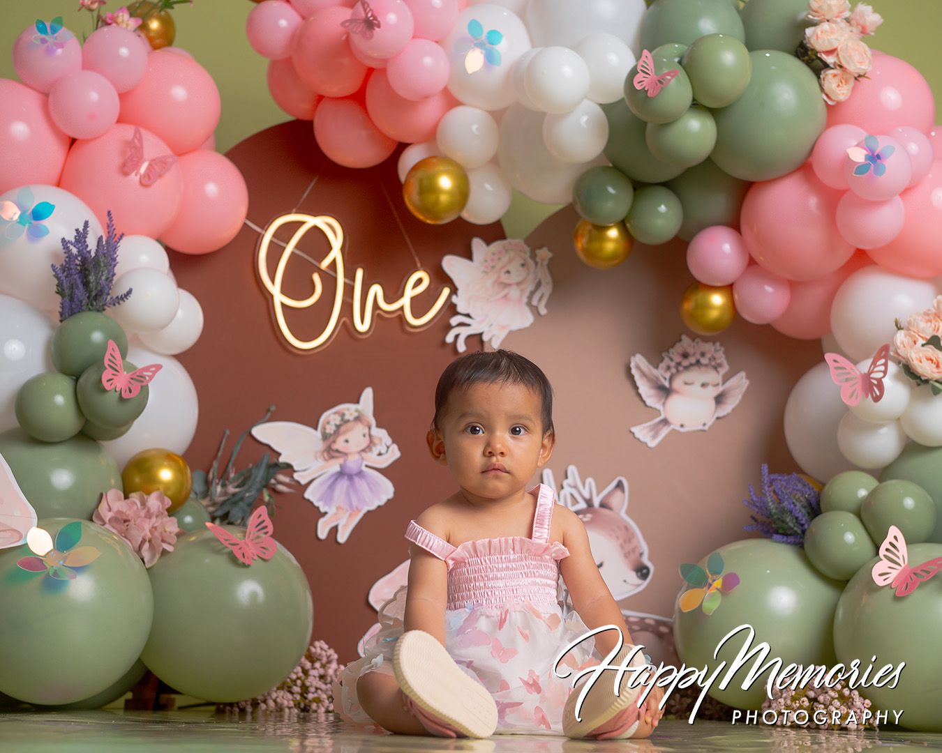 Baby girl sits before a fairy-themed first birthday backdrop with pink, green, and gold balloons spelling One.