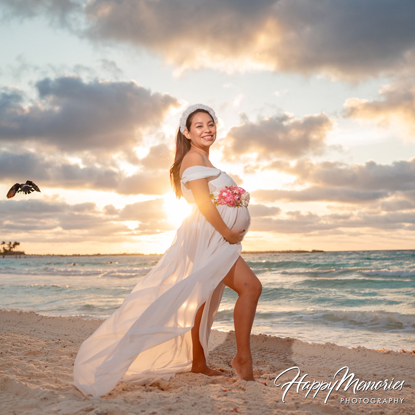 Pregnant woman in white flowing dress poses on beach at golden sunset holding pink flowers.