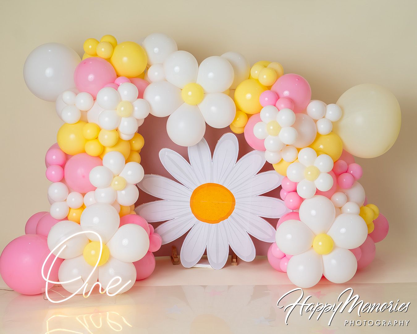 Decorative balloon arch with pink, yellow and white balloons featuring large white daisy flowers against beige wall.
