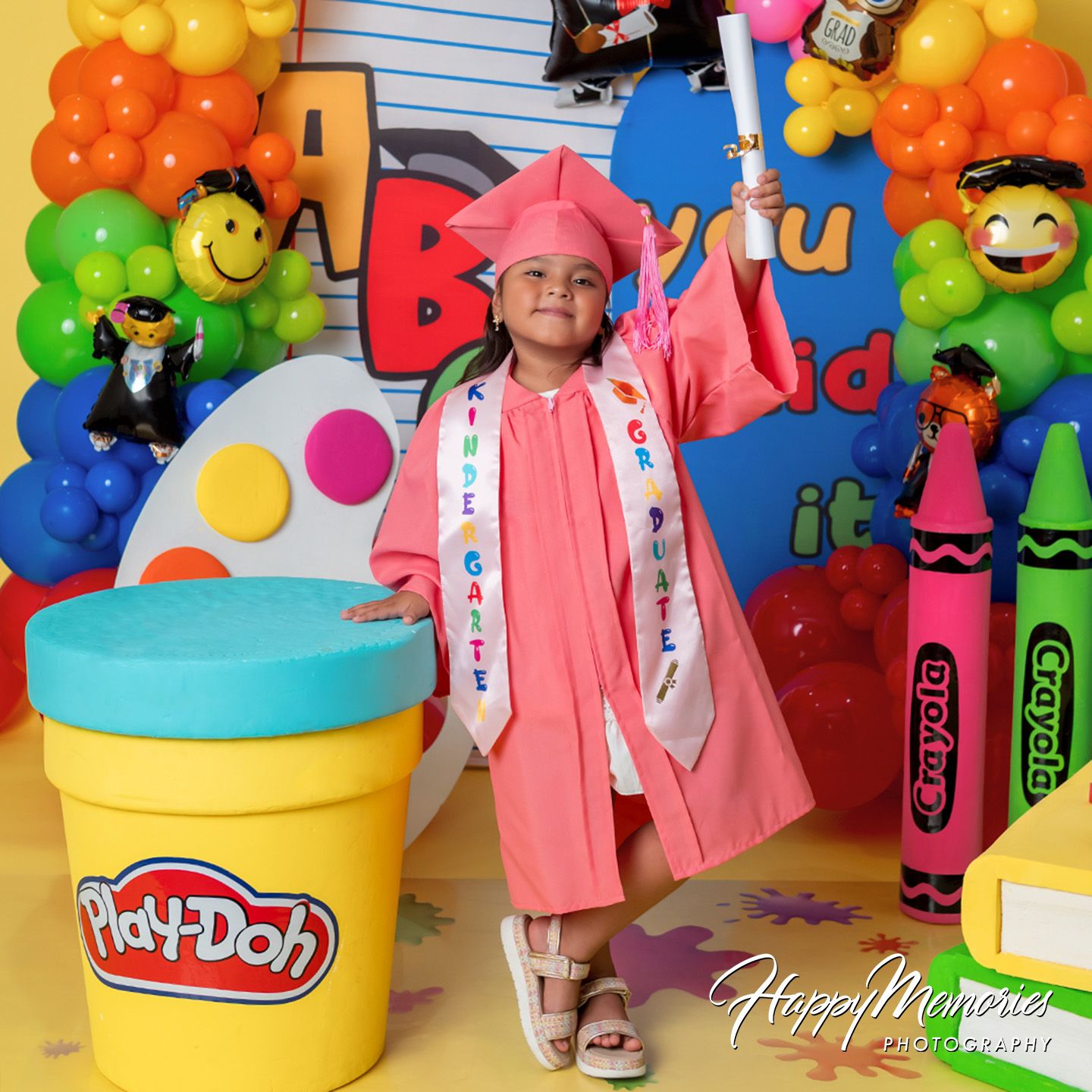 Young girl in pink cap and gown poses at colorful kindergarten graduation backdrop with Play-Doh and Crayola decorations.