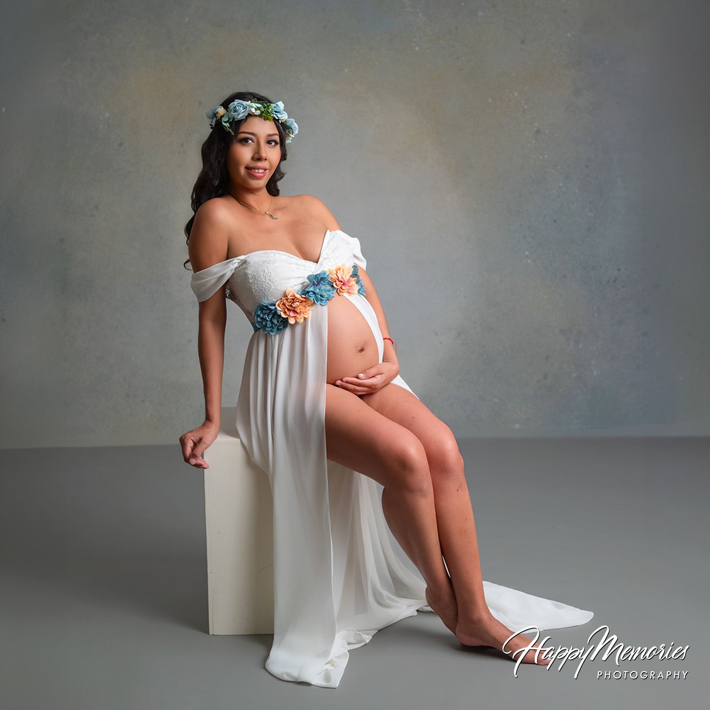 Pregnant woman in white floral gown and flower crown poses elegantly for maternity photography session.