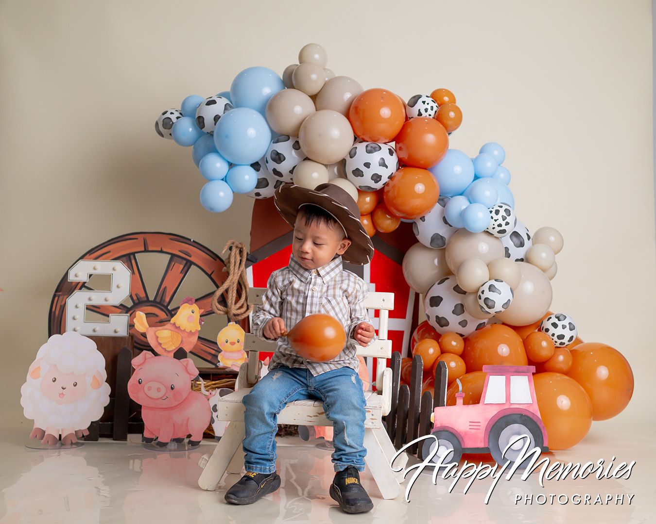 Toddler boy in cowboy hat sits at farm-themed first birthday smash cake setup with colorful balloons.