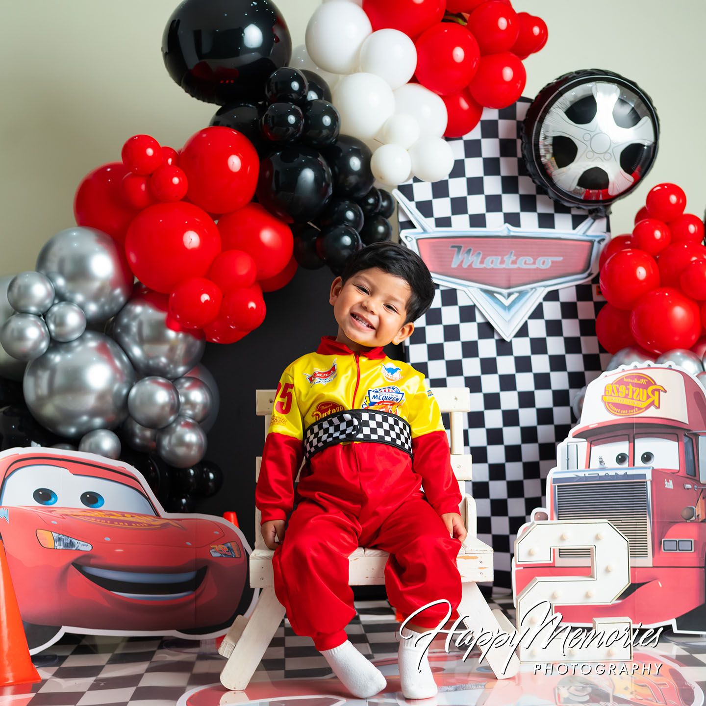 Toddler in Lightning McQueen racing suit poses with Cars-themed balloon arch and character cutouts at birthday party.