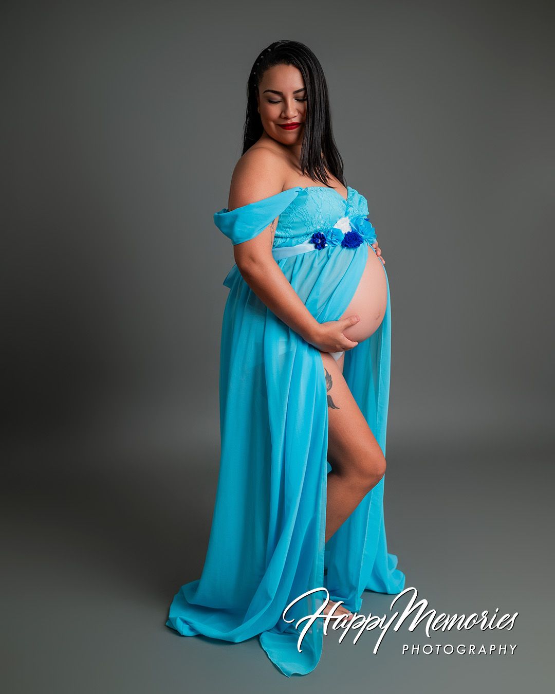 Expectant mother in flowing turquoise off-shoulder gown with floral details during maternity photoshoot.