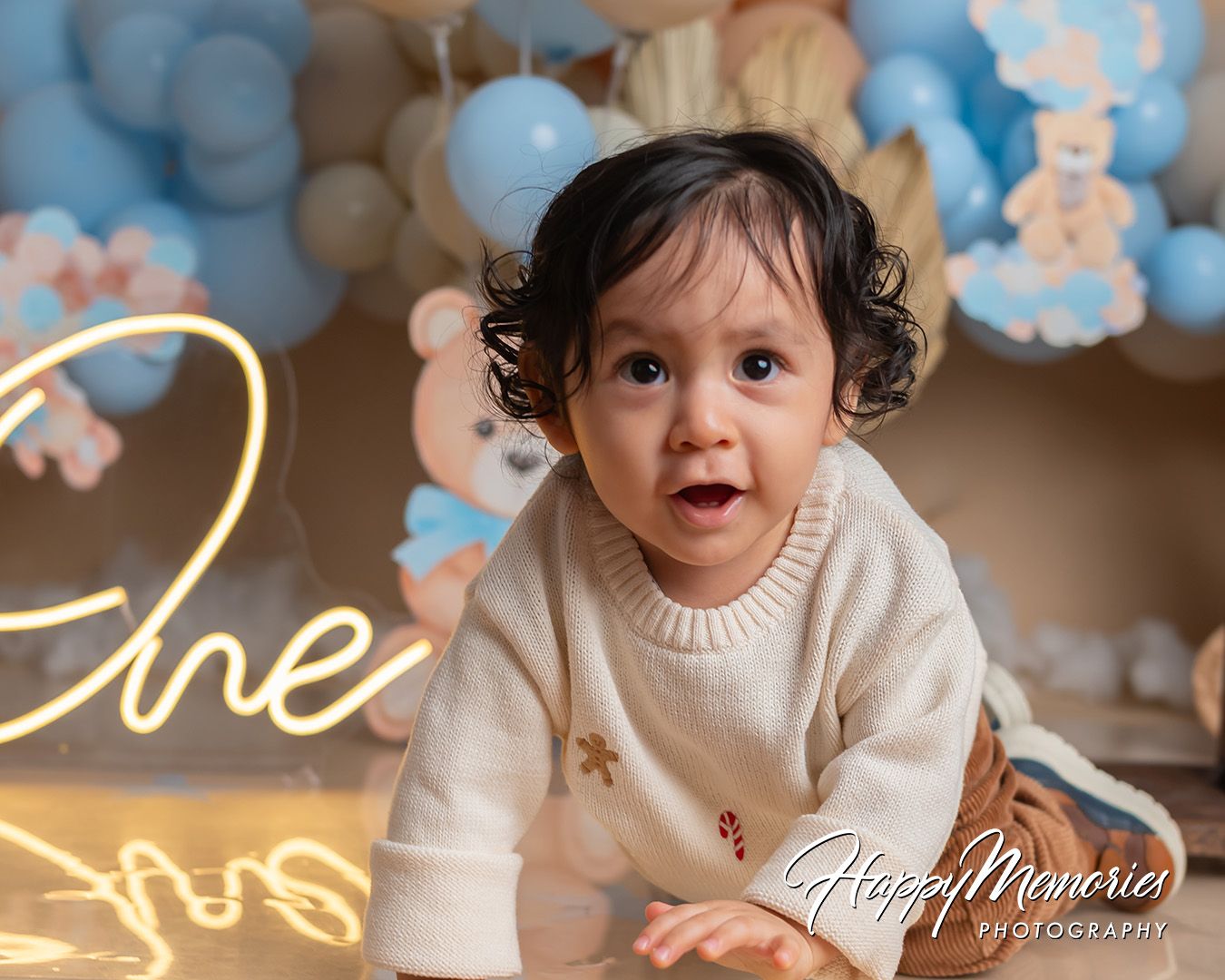 Baby in cream sweater smiles during photo session with blue balloon decor and neon 'One' sign in background.