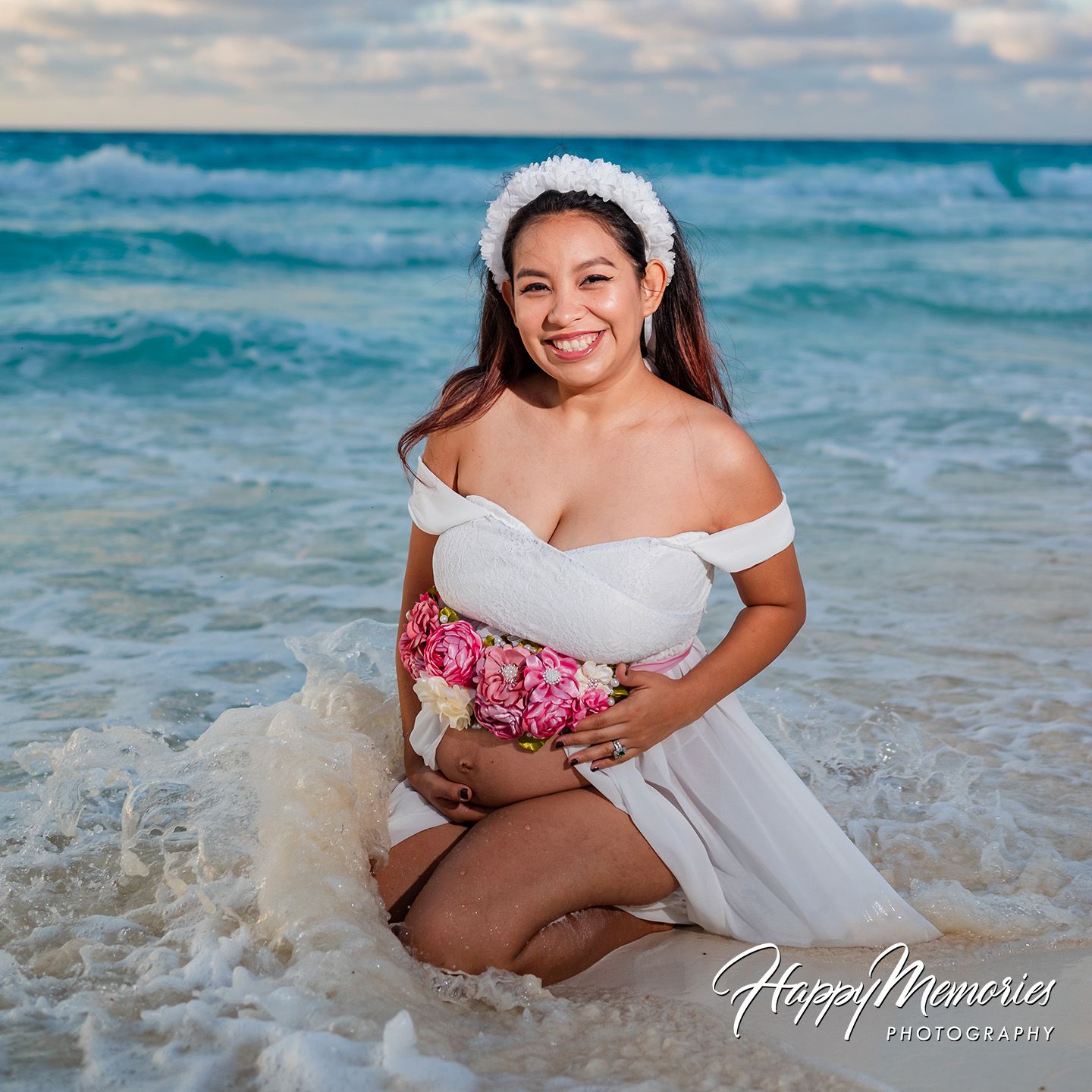 Smiling pregnant woman kneeling in ocean waves wearing white off-shoulder gown with floral bouquet during beach maternity photo session.