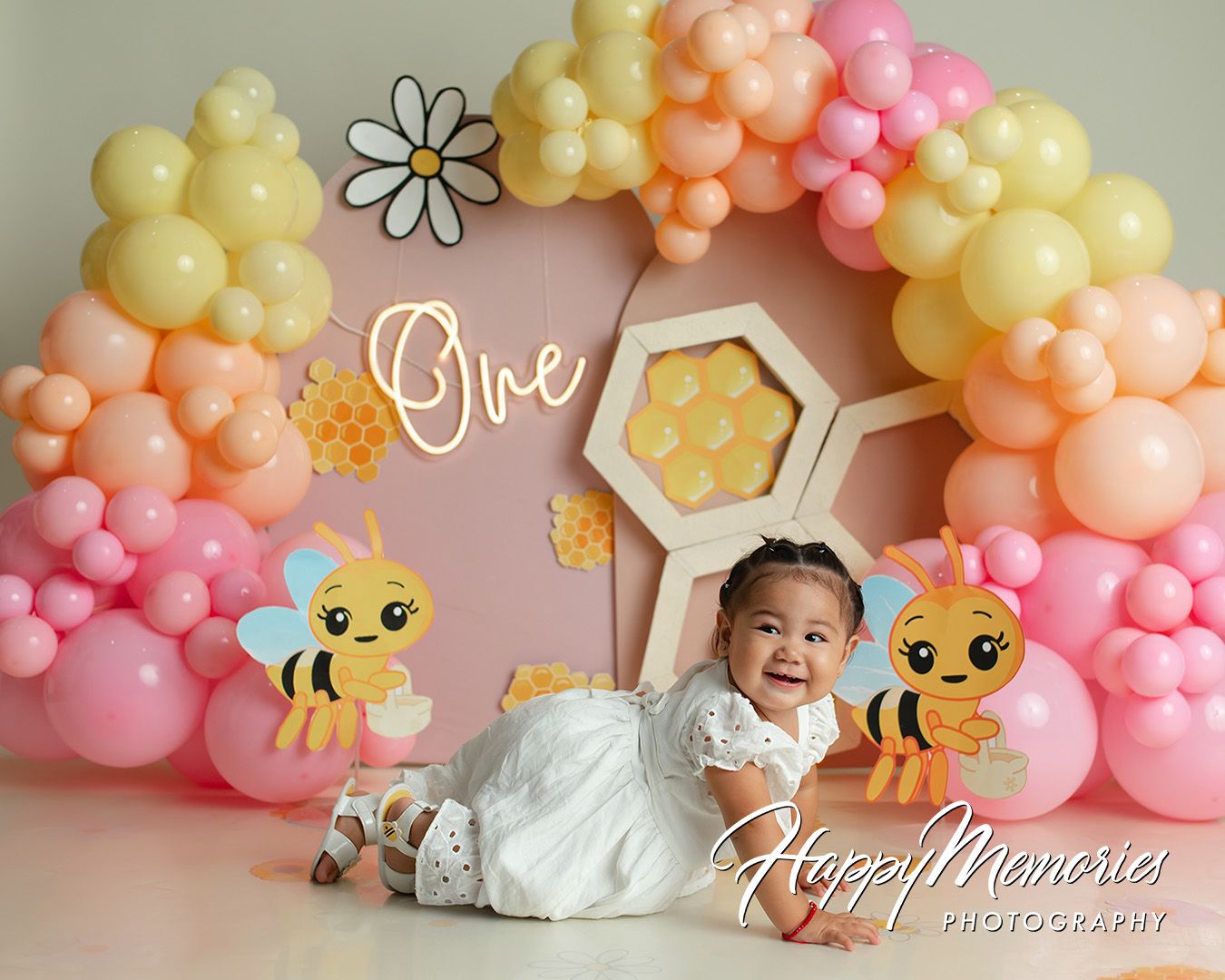 Baby girl smiles at her first birthday party with bee theme balloons, honeycomb backdrop, and colorful arch.
