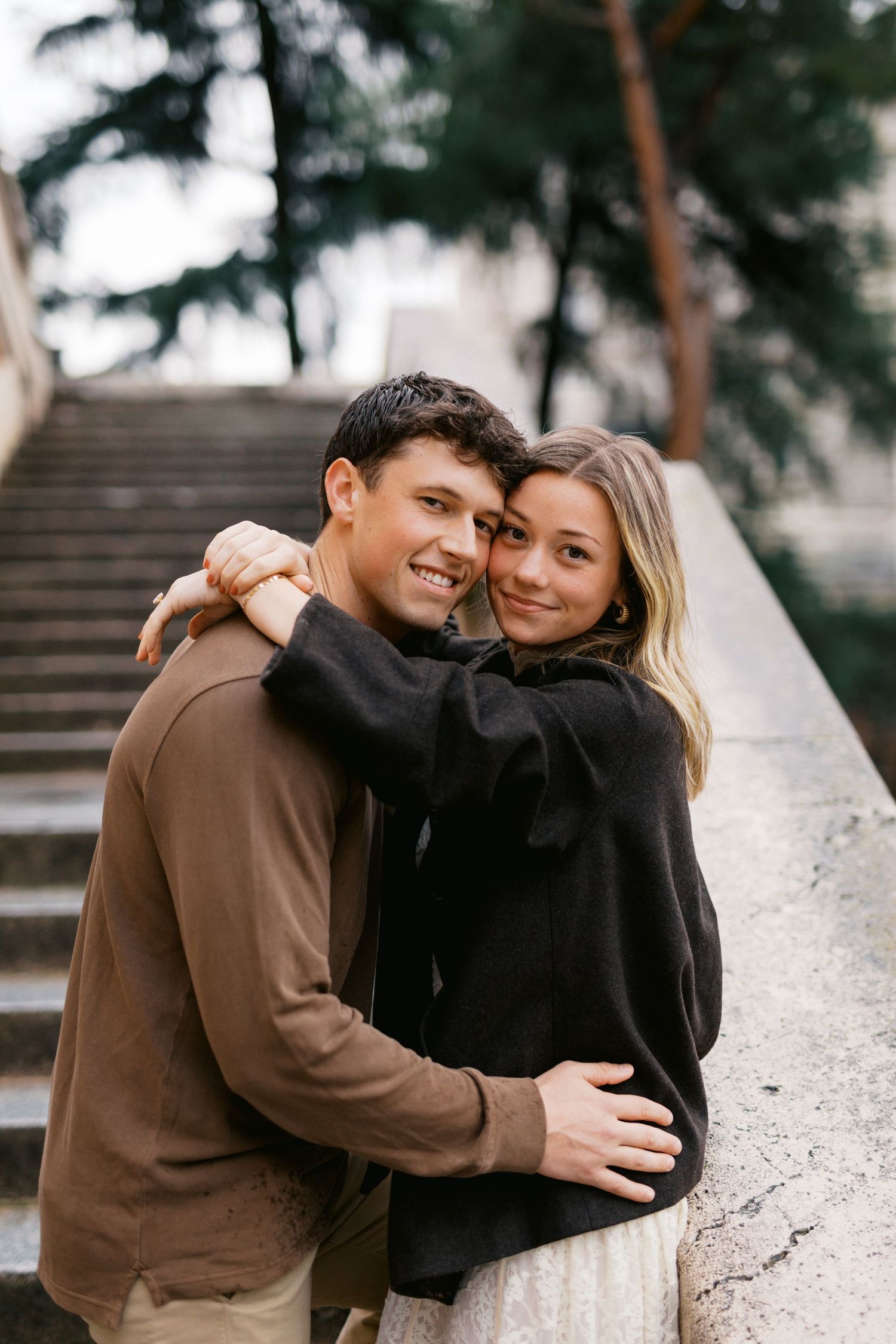 A couple shares intimate embraces while sitting on outdoor stone steps surrounded by pine trees.
