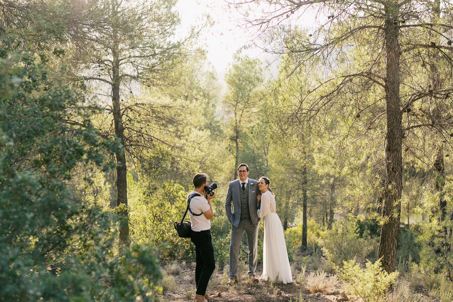 A couple in wedding attire shares an intimate moment in a sunlit forest with golden light filtering through trees.