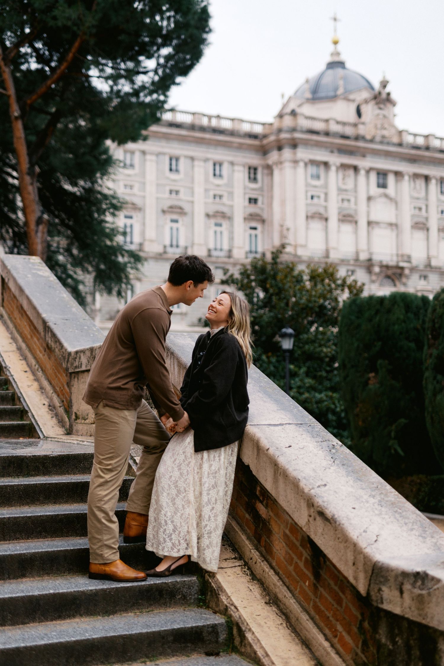 A romantic couple shares tender moments on stone steps with a grand palace building visible in the background.