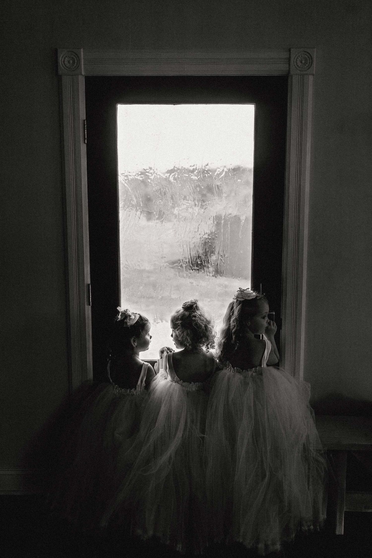 Three ethereal flower girls in white dresses stand silhouetted against a bright window in a dark room.