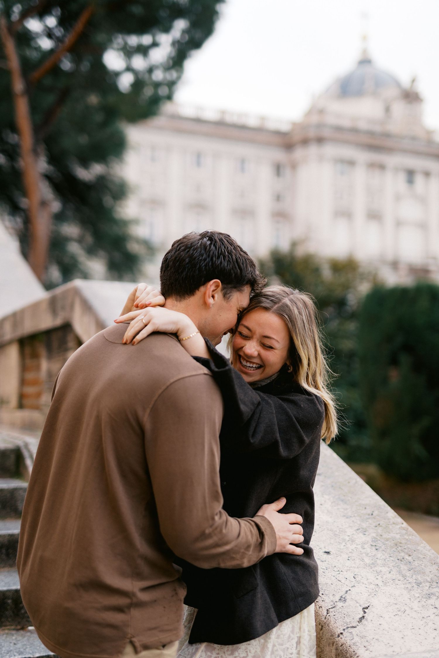 A couple shares a tender embrace while sitting on a stone wall with a grand building visible in the background.
