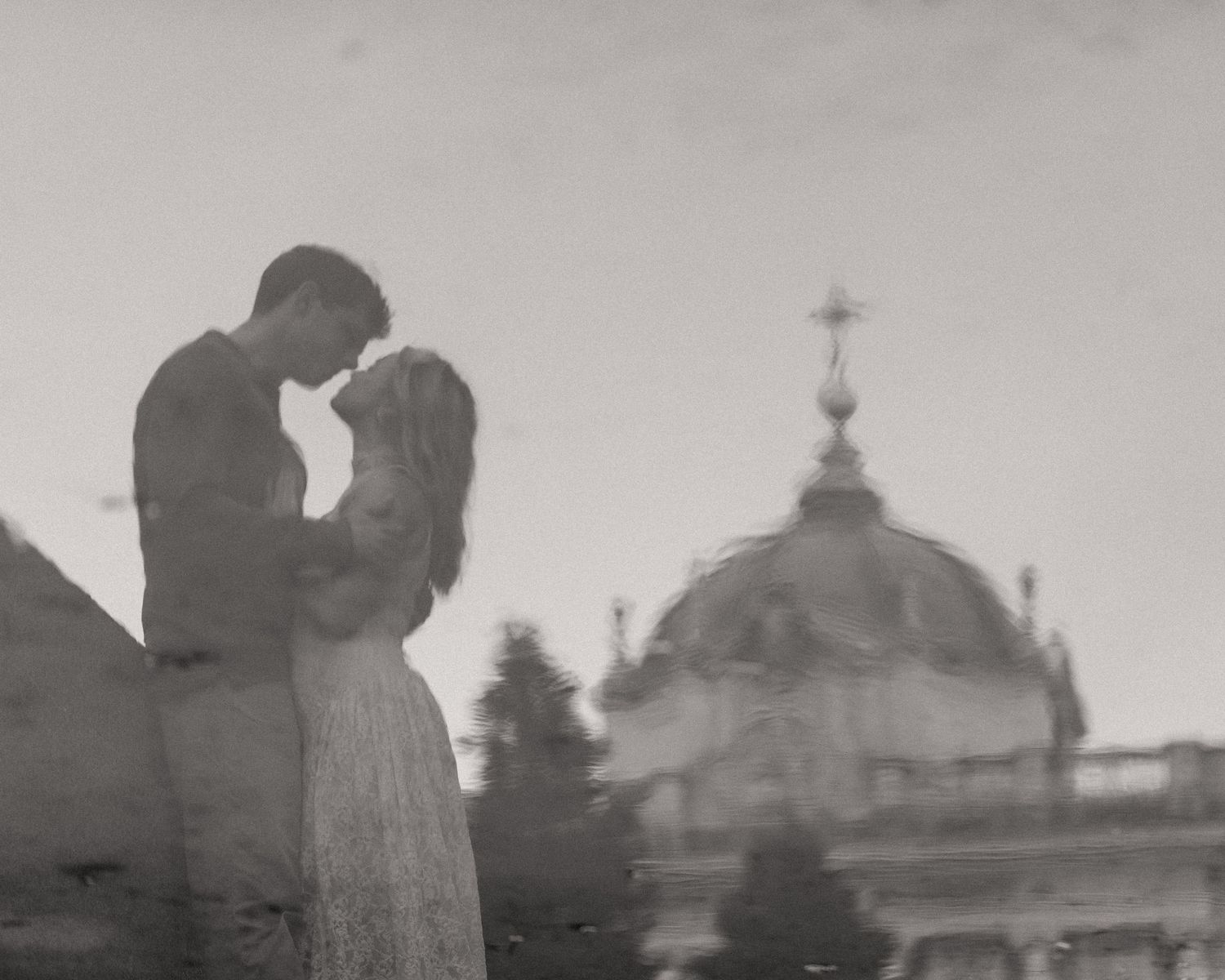 A romantic silhouette of a couple embracing in front of a domed building in a dreamy black and white photograph.
