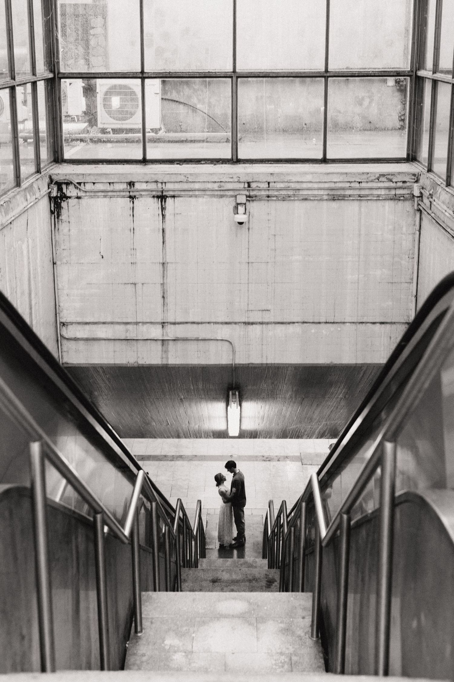 Black and white photo of silhouettes at the top of escalator stairs in a subway station with industrial lighting above.