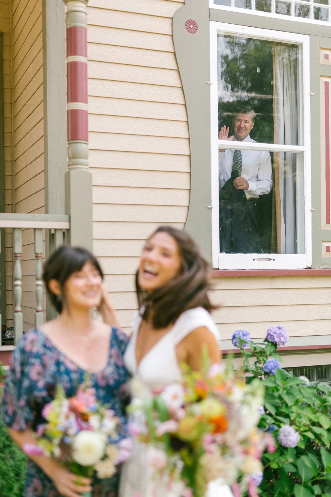Bride and sister share joyful laughter while their father waves from a window behind them during a Marquette wedding.