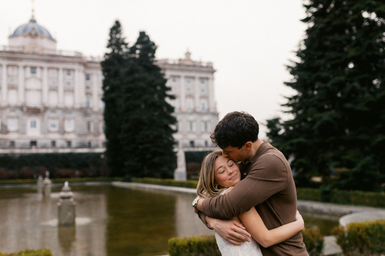 A romantic couple embraces in front of an elegant white palace building with manicured grounds.