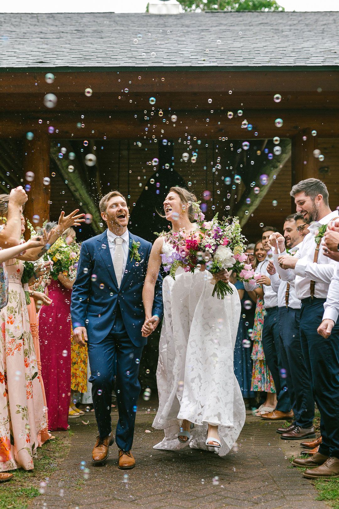 Wedding party celebrates with bubbles outside venue as guests blow soap bubbles in joyful outdoor reception moment.