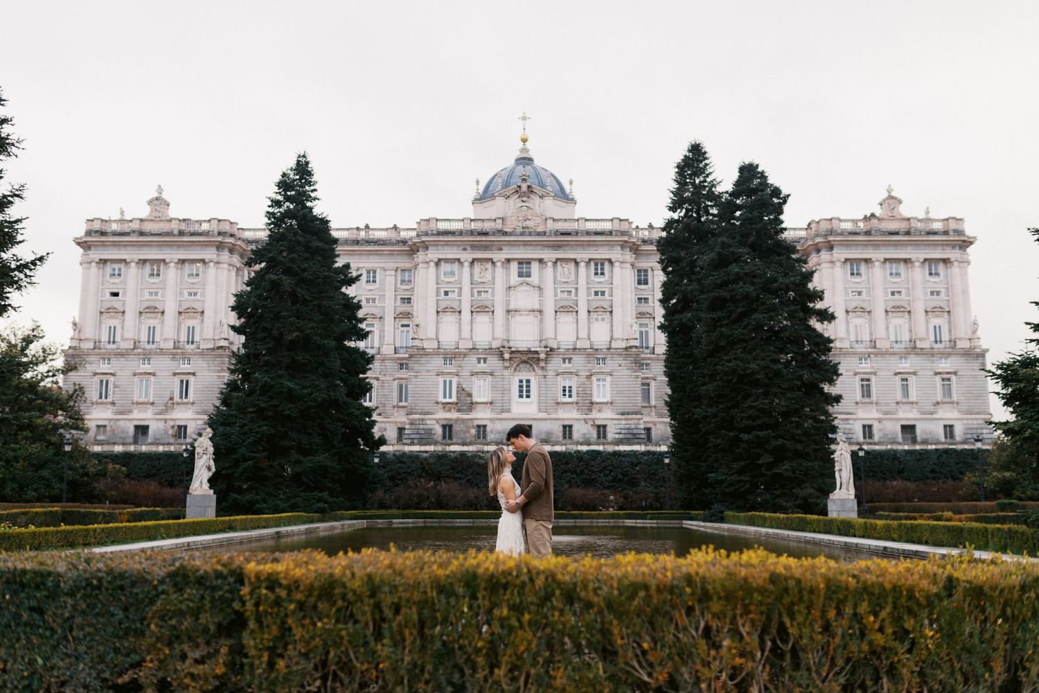 Elegant white palace with classical architecture stands behind a manicured garden with ornate hedges and fountains.