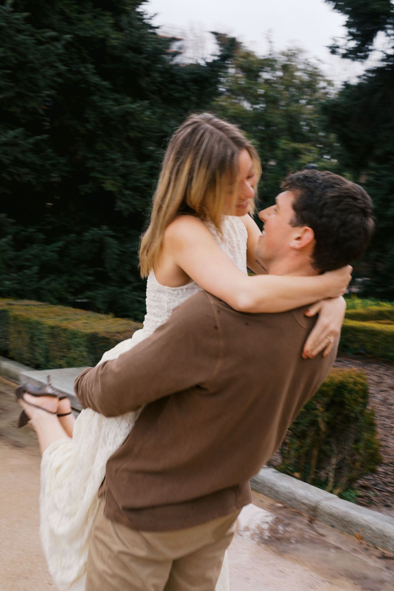 A romantic couple embraces in an outdoor setting with trees and a path visible in the background.