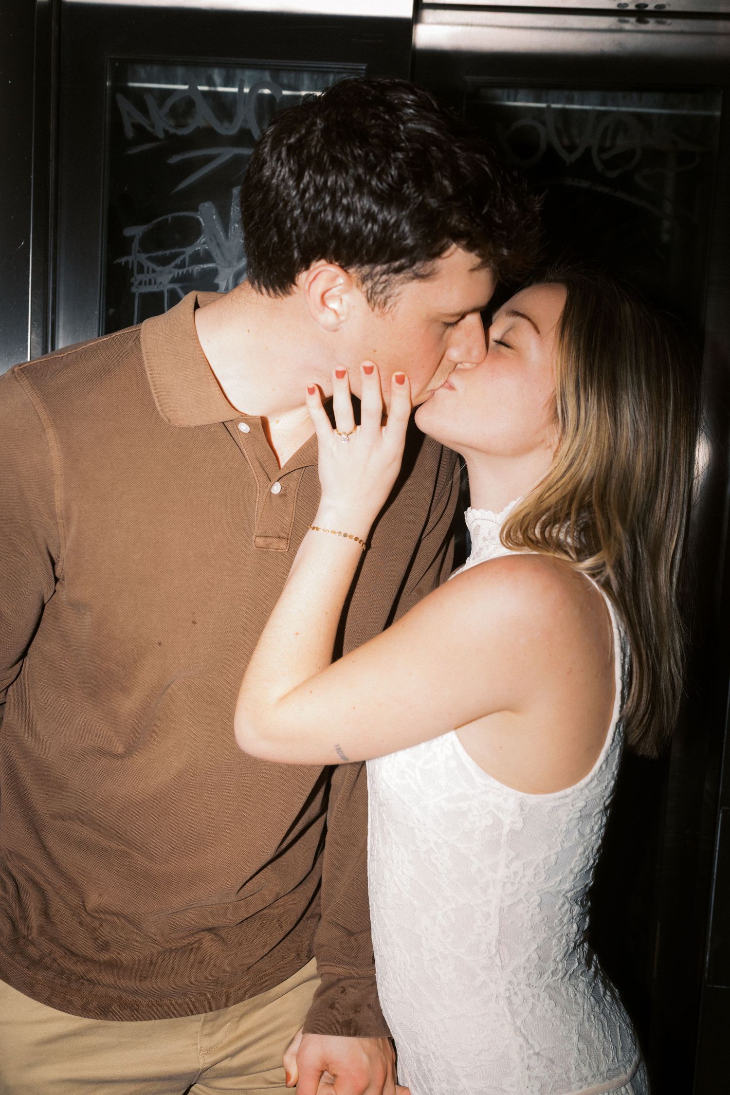 A romantic couple shares an intimate kiss against a dark wall, with one wearing a brown polo and the other in white.