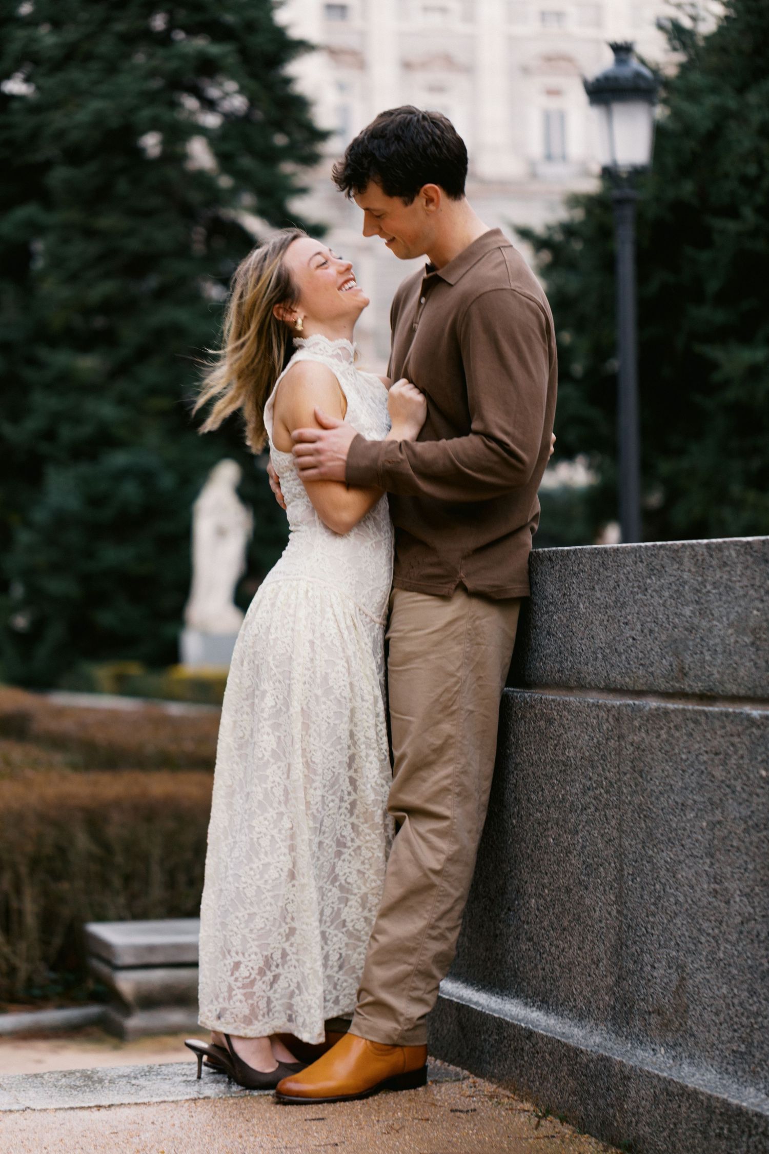 A couple in a romantic embrace on stone steps, with her in a white lace dress and him in a brown suit.