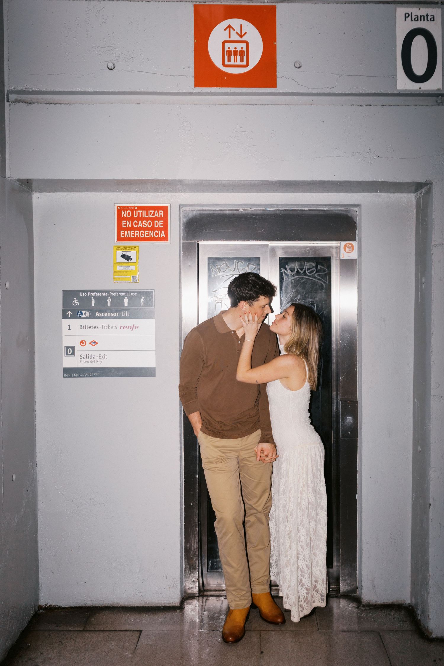 A couple shares an intimate moment while standing in a white elevator doorway with orange signage above.