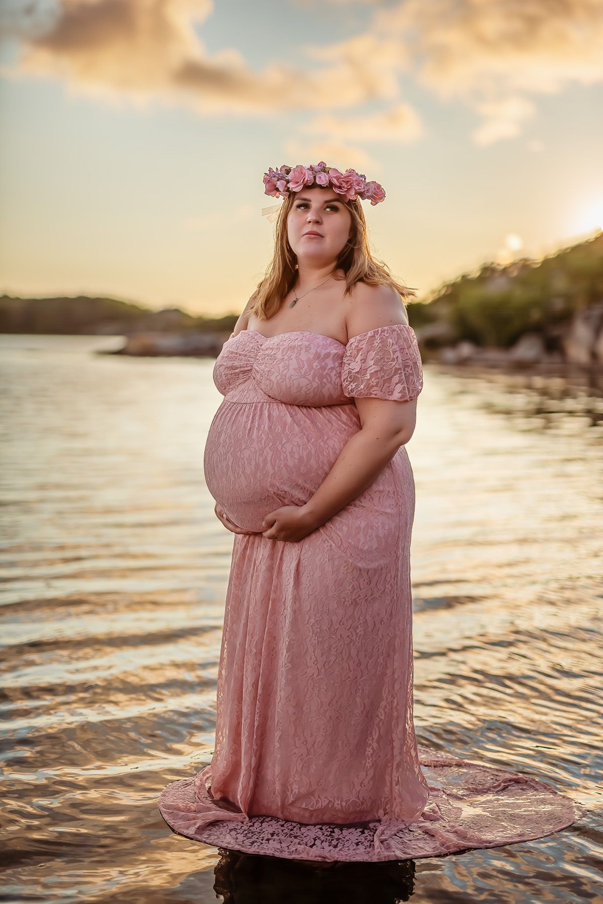 Gravidkvinna i solnedgång i rosa klänning står i vattnet.