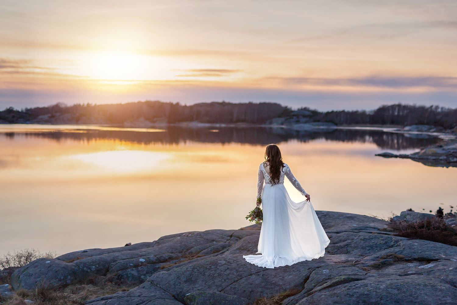 Brud i vacker bröllopsklänning vid solnedgång vid klippor. Håller blombukett. Bröllopsporträtt. Magiskt