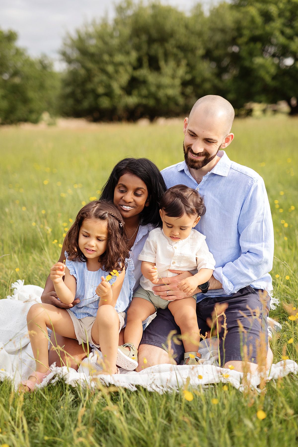 Familjefotografering i naturen. Mamma, pappa och två barn sitter på gräsmattan och tittar på blommorna