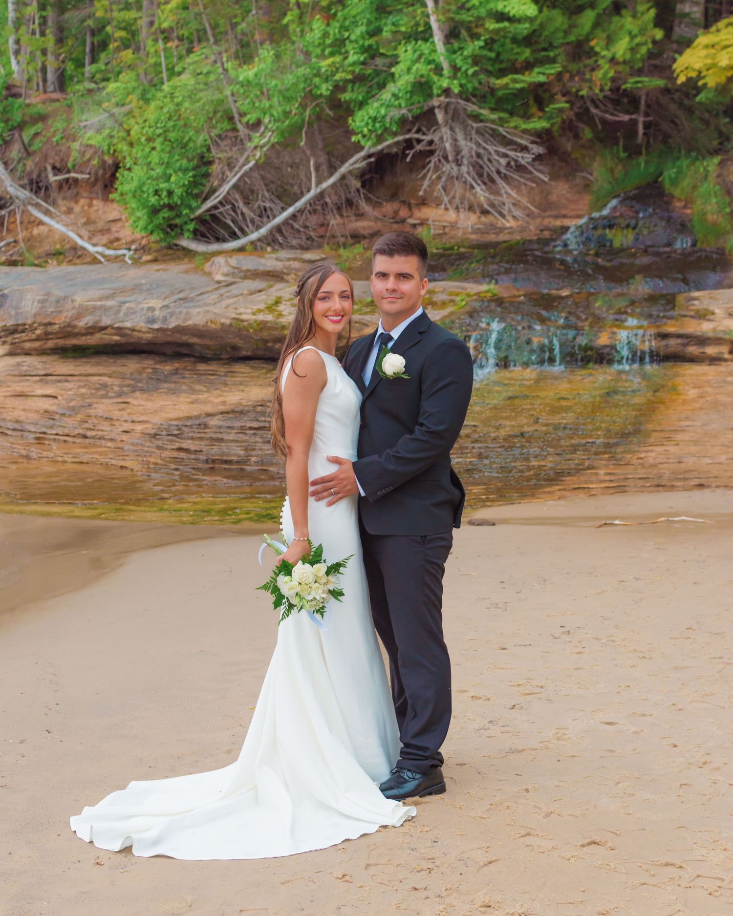 A wedding couple poses on Miners beach near rocky cliffs with exposed tree roots and a small waterfall in the background.
