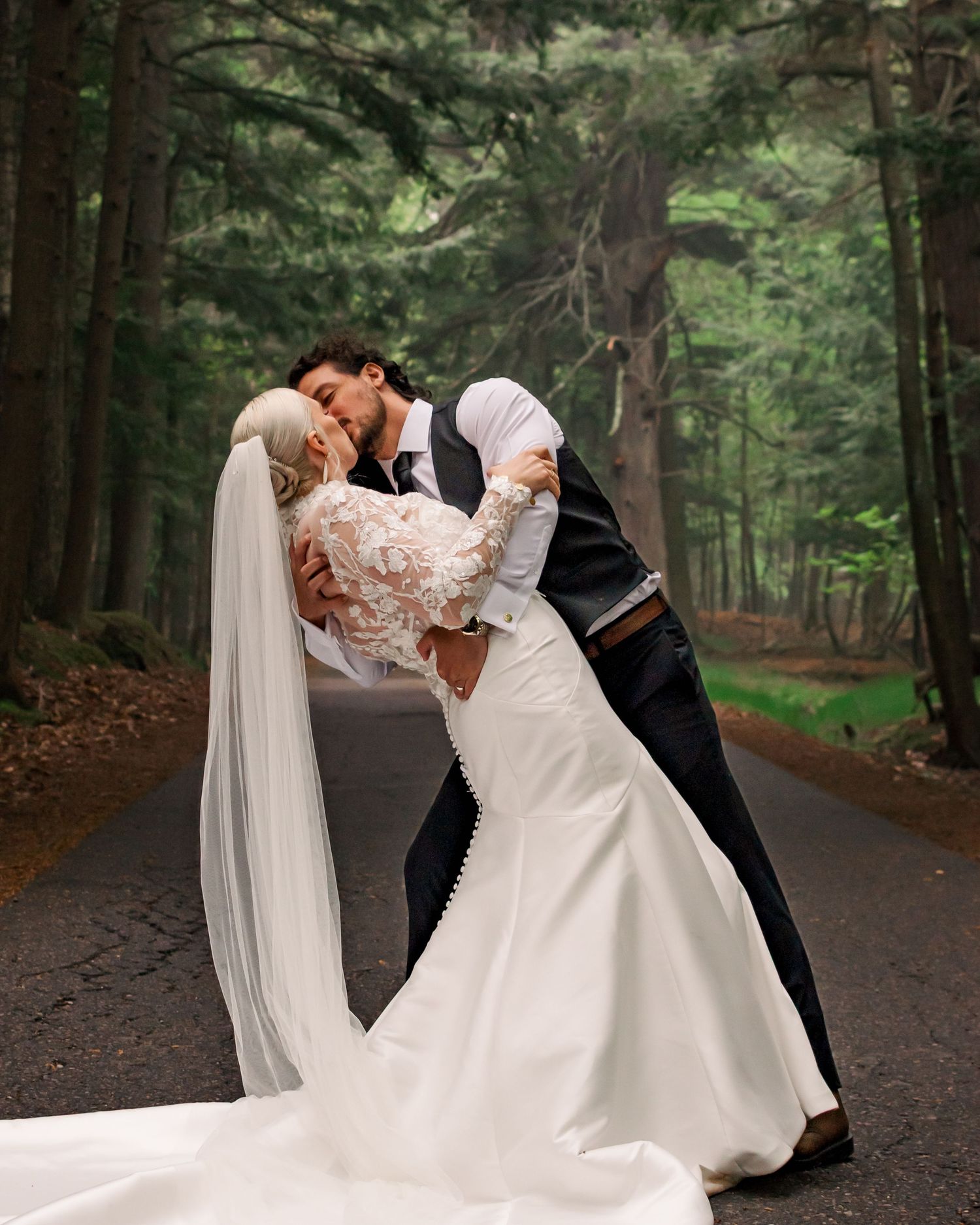 A romantic wedding photo shows a couple in formal attire sharing an intimate dip pose on a wooded forest path in Marquette Michigan