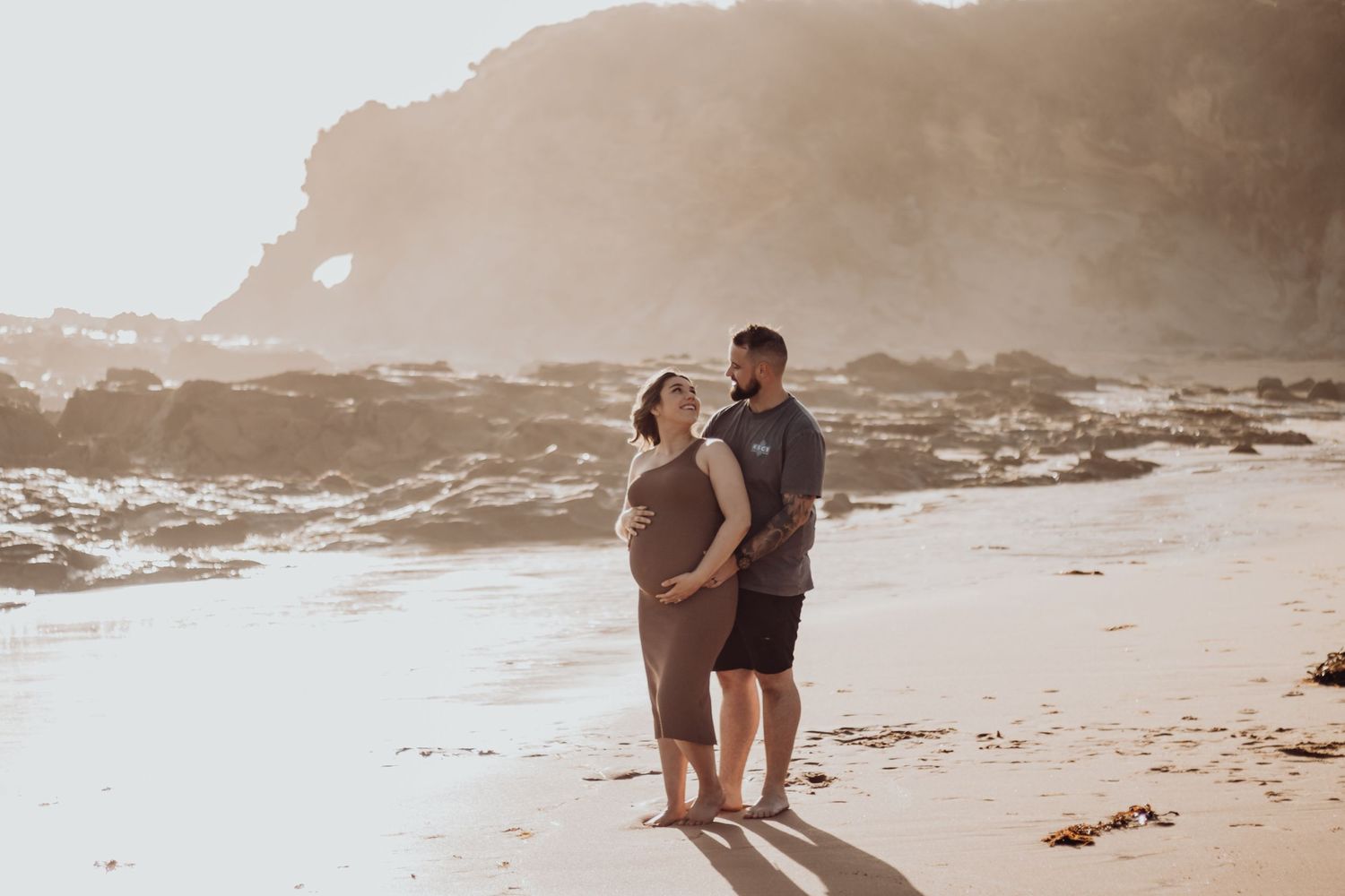 Pregnant woman and husband holding her belly on the beach.