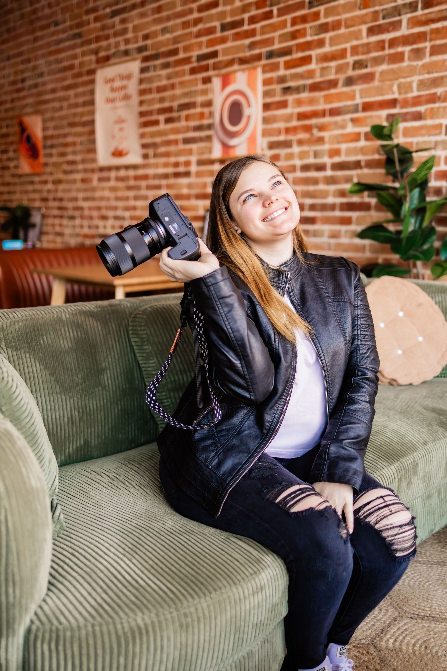 A photographer sits casually on a green couch holding a camera in a cozy space with exposed brick walls.