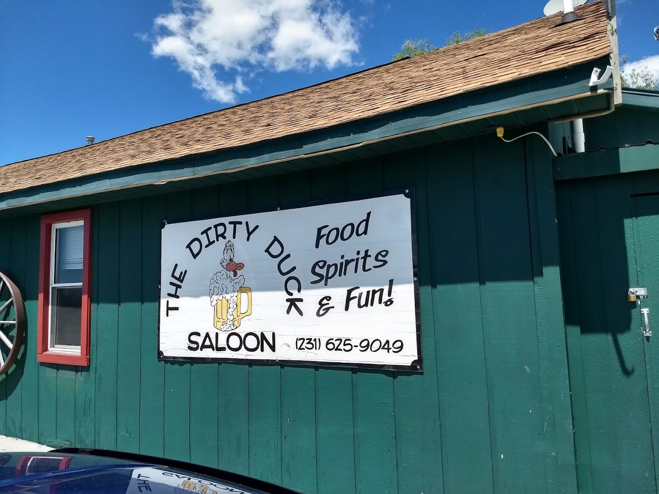 A green-painted building with a wooden roof houses a saloon, featuring signs for food and spirits against a blue sky.