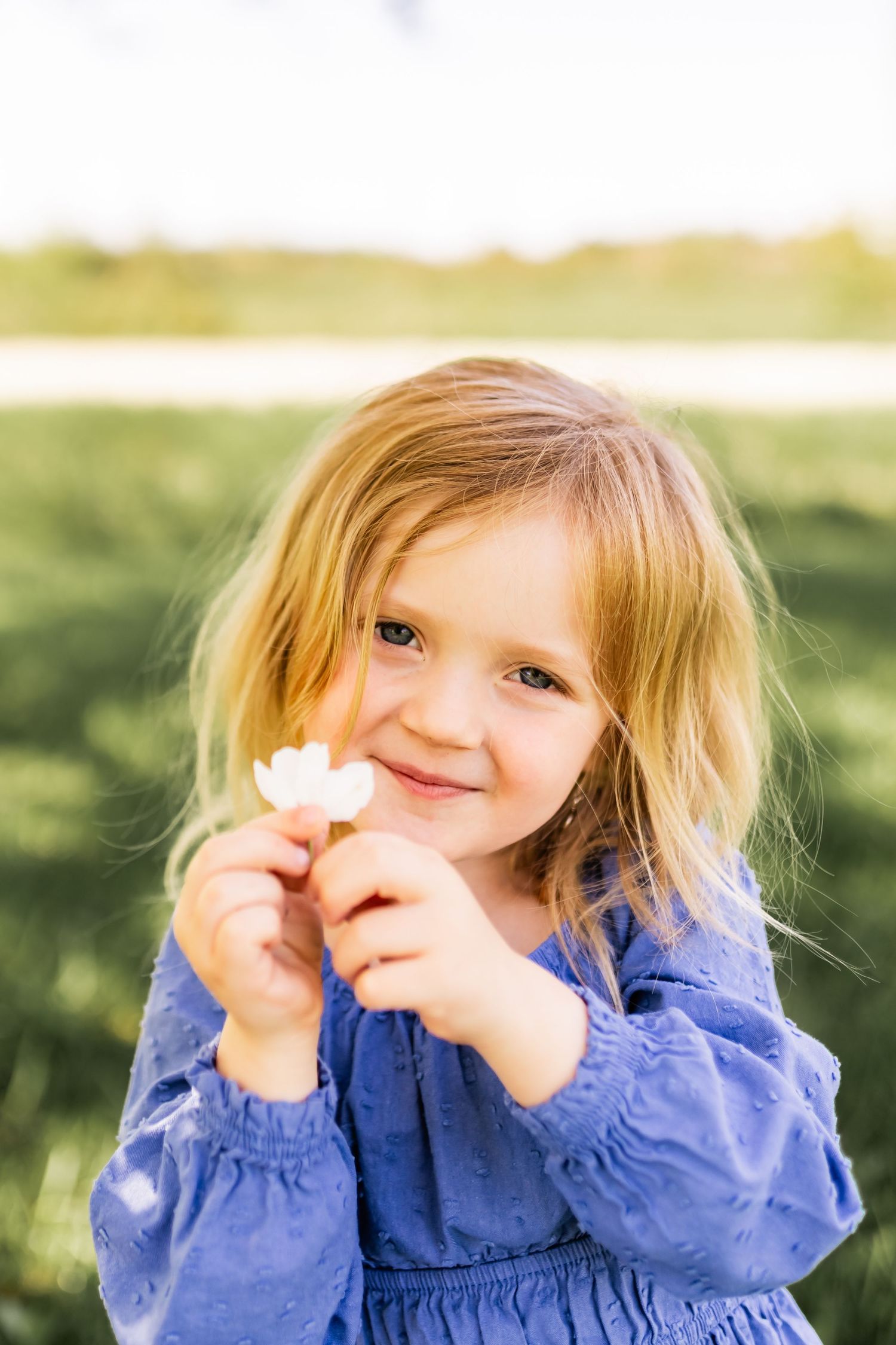 Young child in blue dress holds a small white flower while smiling outdoors on a sunny day.
